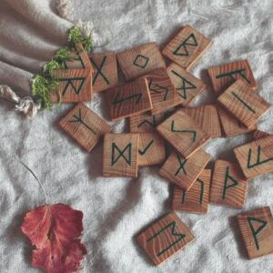 Hand-carved wooden runes resting on soft linen with a red leaf — a quiet reflection of the Elder Futhark’s ancient language of spirit and nature.